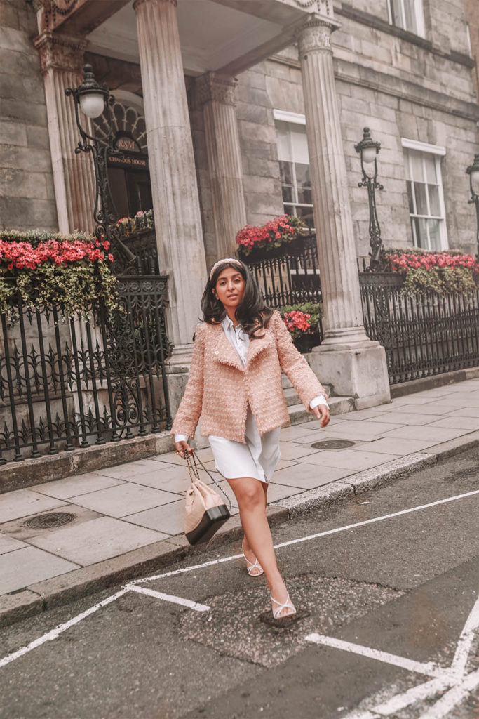 Luxury fashion blogger Shloka Narang of The Silk Sneaker shares how to style a tweed jacket for a luxury Chanel look in a white shirt dress, Chanel backpack, Prada strappy sandals and headband